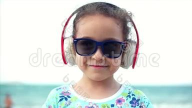 海滩上快乐的小女孩戴着太阳镜，听着耳机上的音乐，看着相机，微笑着，还有一个。